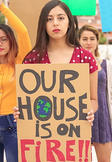 A young woman holding a hand-lettered cardboard sign that says, 'Our house is on fire!!" while looking gravely at the camera. The 'O' in 'house' is filled with a drawing of the Earth.